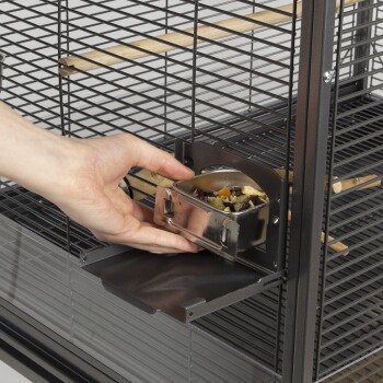 Eine Hand, die eine Edelstahlnapf mit gemischtem Haustierfutter in einen Metallkäfig mit Holzstangen und einer herausziehbaren Schublade stellt.