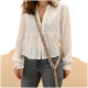 Eine Frau trägt eine weiße bestickte Bluse und graue Jeans, hält ein gemustertes Band mit einem Clip. Der Hintergrund ist ein sanftes Beige.