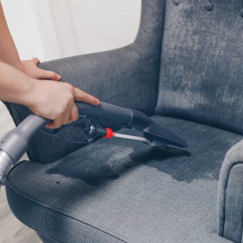 Eine Person benutzt einen Staubsaugeraufsatz, um einen nassen grauen gepolsterten Stuhl zu reinigen, mit sichtbaren Wasserflecken auf dem Stoff.