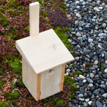 Holzvogelhaus mit schrägem Dach, auf moosigem Boden umgeben von Kieselsteinen platziert.