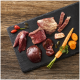 Verschiedene rohe Fleisch- und Innereienstücke mit Karotte und Samen auf einem schwarzen Schiefer‑Servierbrett arrangiert.
