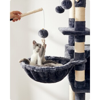 Eine verspielte grau-weiße Katze greift nach einem hängenden Pom-Pom-Spielzeug, während sie in einer gemütlichen, dunkelgrauen Hängematte, die an einem Kratzbaum befestigt ist, liegt.