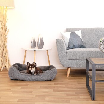 Ein kleiner braun-weißer Hund entspannt sich in einem gemütlichen grauen Haustierbett auf einem Holzboden, mit einem modernen grauen Sofa und dekorativen Gegenständen im Hintergrund.