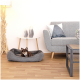 Ein kleiner braun-weißer Hund entspannt sich in einem gemütlichen grauen Haustierbett auf einem Holzboden, mit einem modernen grauen Sofa und dekorativen Gegenständen im Hintergrund.