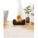 Ein flauschiger orangefarbener Pomeranian sitzt glücklich in einem schwarzen Haustierbett, umgeben von einem lichtdurchfluteten Raum mit einem grauen Sofa und einer Topfpflanze.