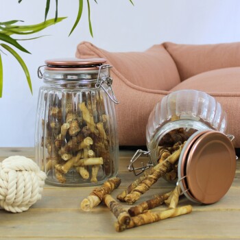 Ein Glasbehälter voller Hundekauartikel steht auf einem Holztisch, mit einigen Kauartikeln, die herumliegen. Im Hintergrund befindet sich ein weiches rosa Haustierbett.