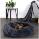 Ein schwarz-brauner Hund ruht in einem plüschigen, runden, dunkelgrauen Hundebett auf einem gefliesten Boden, mit einem kleinen Holztisch und einer Topfpflanze in der Nähe.