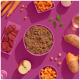 Ein lebendiges Flatlay von Haustierfutterzutaten auf einem rosa Hintergrund, mit einer Schüssel mit Nassfutter für Hunde, gewürfeltem Fleisch, Süßkartoffeln, Karotten und einem Apfel.