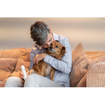 Ein Mann mit grauen Haaren umarmt sanft einen kleinen braunen Hund, während er auf einem plüschigen Sofa sitzt und ein Haustierpflegeprodukt in der Hand hält.