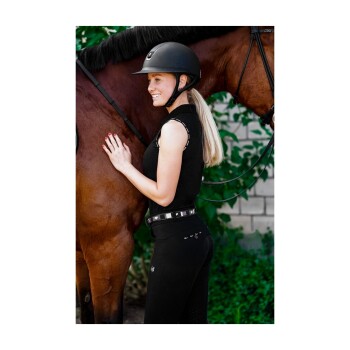 Eine Frau in einem schwarzen Reitoutfit und Helm steht neben einem braunen Pferd und berührt sanft seine Seite, mit Grün im Hintergrund.