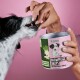 Eine Person füttert einen schwarz-weißen Hund mit einem Leckerli aus einem grün-pinken Behälter mit der Aufschrift "Darmpflege Drops" vor einem pinken Hintergrund.