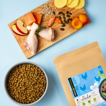 Ein Holzschneidebrett zeigt rohe Hähnchenschenkel, Lachs, Apfelscheiben, Aprikosen und Samen, mit Tierfutter in der Nähe.