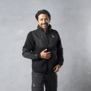 Eine Person trägt eine schwarze Fleecejacke mit Reißverschluss und einem Logo auf der linken Brust, steht vor einem grauen Hintergrund.