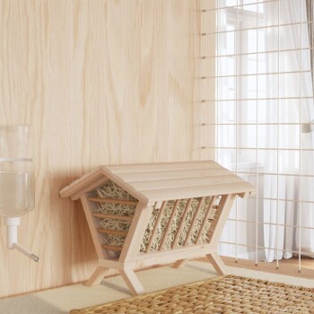 Holz-Haustierhaus mit Heu innen, neben einem Wasserspender, vor einer hellen Holzwand und einer Gittertrennwand.