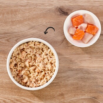 Eine Schüssel mit feuchtem Katzenfutter mit zerrissenem Fleisch steht auf einer Holzoberfläche, neben einer kleinen Schüssel mit bunten rohen Fischstücken.