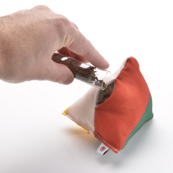 Eine Hand, die braunes Haustierfutter aus einem Glasrohr in einen bunten, dreieckigen Stoffbeutel mit orangefarbenen, grünen und gelben Abschnitten gießt.