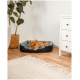 Ein kleiner, flauschiger Hund ruht sich in einem gemütlichen, grau-schwarzen Hundebett auf einem Holzboden aus, mit einer Topfpflanze und einer weißen Kommode in der Nähe.