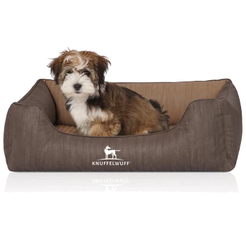 Ein kleiner, flauschiger Hund sitzt bequem in einem stilvollen braunen Haustierbett mit geripptem Design, das das Logo "KNUFFELWUFF" trägt.