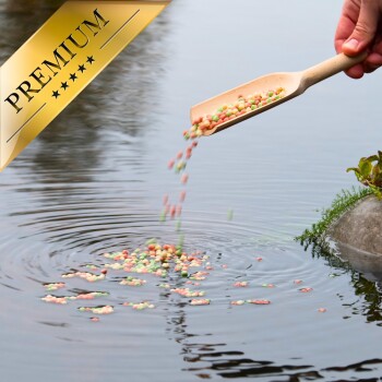Eine Hand gießt bunte Fischfutterpellets aus einer Holzschaufel in einen ruhigen Teich, mit einem goldenen Banner "PREMIUM" in der Ecke.