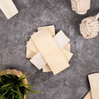 Ein Haufen beiger Hundekauartikel, die auf einer grauen Oberfläche angeordnet sind, mit einem geflochtenen Korb aus grünem Gras und zwei Seilspielzeugen in der Nähe.