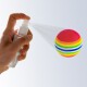 Eine Hand hält eine Sprühflasche, die einen bunten, gestreiften Haustier-Spielzeugball besprüht, der lebendige pinke, gelbe, grüne und blaue Farben zeigt.