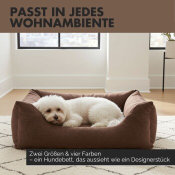 Weißer flauschiger Hund in braunem Designer‑Hundebett in modernem Raum; deutscher Text: 'PASST IN JEDES WOHNAMBIENTE' und 'Zwei Größen & vier Farben'.