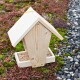 Holz-Vogelfutterhaus mit einem schrägen Dach und einer Schale, die mit Vogelfutter gefüllt ist, auf einem moosigen Boden mit Kieselsteinen.