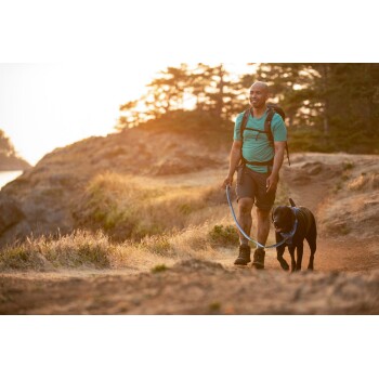 Eine Person in einem türkisfarbenen Shirt und Shorts führt einen schwarzen Hund an der Leine auf einem Schotterweg, umgeben von Gras und Bäumen bei Sonnenuntergang.