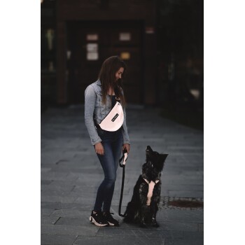 Eine Person in einer Jeansjacke und Jeans hält eine Leine für einen schwarzen Hund im Geschirr, mit einer pinken "PARKA"-Tasche über der Schulter.