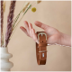 Eine Hand hält ein braunes Leder-Haustierhalsband mit einer silbernen Schnalle, das vor einem weichen beigen Hintergrund mit dekorativen, getrockneten Blumen präsentiert wird.