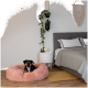 Ein schwarz-brauner Hund entspannt sich in einem rosa Haustierbett auf einem Holzboden in der Nähe eines grauen Bettes und eines kleinen Beistelltisches mit Pflanzen und einer Kerze.