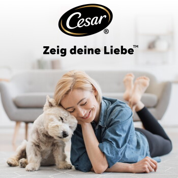 Cesar-Logo und Text 'Zeig deine Liebe' über einer lächelnden Frau, die auf dem Boden liegt und einen kleinen weißen Hund im Wohnzimmer kuschelt.