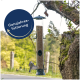 Ein Vogelhaus, das von einem Baumast hängt, gefüllt mit Samen, mit einem kleinen Vogel, der in der Nähe fliegt. Textüberlagerung lautet "Ganzjahresfütterung."