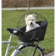 Ein kleiner weißer Hund sitzt in einem schwarzen Fahrradkorb mit einem Drahtkäfig oben, umgeben von grünem Gras und einer Straße im Hintergrund.