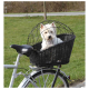 Ein kleiner weißer Hund sitzt in einem schwarzen Fahrradkorb mit einem Drahtkäfig oben, umgeben von grünem Gras und einer Straße im Hintergrund.