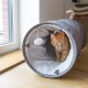 Eine orangefarbene getigerte Katze, die aus einem grauen Stofftunnel mit weichem weißen Innenraum hervorlugt, der ein hängendes Pom-Pom-Spielzeug hat.