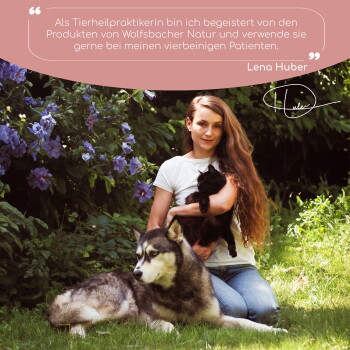 Eine Frau mit einer schwarzen Katze sitzt neben einem wolfähnlichen Hund in Grün und lila Blumen. Text: "Als Tierheilpraktikerin..." - Lena Huber.