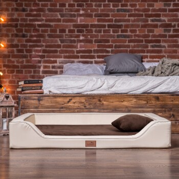 Ein stilvolles, cremefarbenes Haustierbett mit einem braunen Kissen, auf einem Holzboden vor einer Ziegelwand platziert, mit einem gemütlichen Bett im Hintergrund.