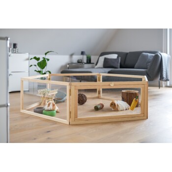 Un enclos en bois pour animaux de compagnie avec un cochon d'Inde à l'intérieur, entouré de jouets et de nourriture, situé dans un salon moderne avec un canapé et des plantes.