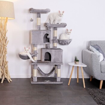 Drei weiße Katzen spielen und entspannen auf einem mehrstöckigen grauen Katzenbaum mit Hängematten und Kratzpfosten, neben einem modernen grauen Sofa und einer Lampe.