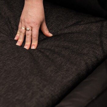 Ein braunes Hundebett mit einem plüschigen Design, mit einem grauen Hund, der bequem darauf sitzt, vor einer hellen Wand.