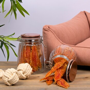 Zwei Glasbehälter gefüllt mit orangefarbenen Haustierleckereien stehen auf einem Holztisch neben einem rosa Haustiersofa und einem weißen Seilspielzeug.