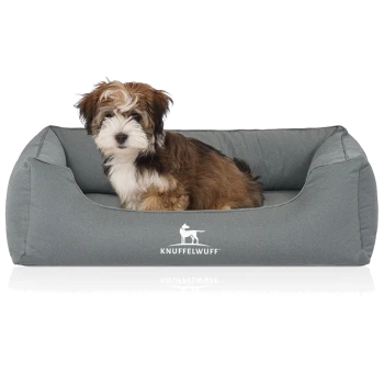 Flauschiger braun-weißer Welpe sitzt in einem grauen Haustierbett mit weißem 'KNUFFELWUFF'-Logo.