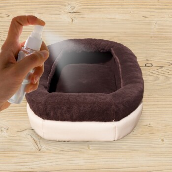 Eine Hand sprüht ein Haustierbett mit einem Nebel aus einer klaren Flasche, das ein gemütliches braunes und cremefarbenes Haustierbett auf einer Holzoberfläche zeigt.