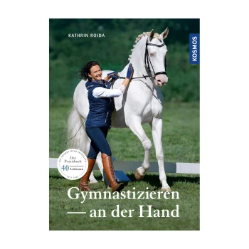 Eine Frau in einer marineblauen Weste führt ein weißes Pferd auf einer grünen Wiese, mit dem Buch 'Gymnastizieren — an der Hand' von Kathrin Roida sichtbar.