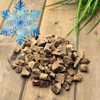 Haufen gefriergetrockneter brauner Futterwürfel auf einem Holzbrett, oben rechts eine Pflanze, blaues Schneeflocken-Symbol mit der Aufschrift ‚Gefrier-getrocknet‘.