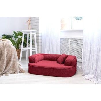 Ein gemütliches rotes Haustierbett mit runder Form und zwei seitlichen Kissen, in einem hellen Raum mit weißen Vorhängen und grünen Pflanzen platziert.