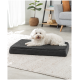 Ein flauschiger weißer Hund ruht auf einem schwarzen Haustierbett mit weichem Oberteil, das auf einem hellen Teppich in einem modernen Wohnzimmer platziert ist.