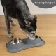 Ein schwarz-grauer Hund frisst aus einem doppelten Edelstahlnapf, der auf einer grauen, pfotenförmigen Matte steht, mit dem Text "Nutzungsbeispiel" in der Ecke.