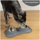 Ein schwarz-grauer Hund frisst aus einem doppelten Edelstahlnapf, der auf einer grauen, pfotenförmigen Matte steht, mit dem Text "Nutzungsbeispiel" in der Ecke.